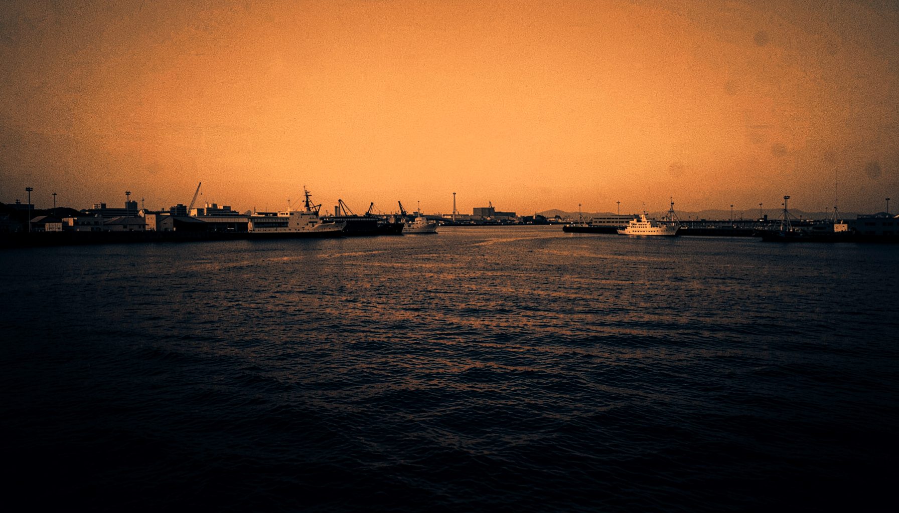 A wide cinematic duotone of Yokosuka harbor at dusk, deep navy water meeting warm amber sky, moody and atmospheric